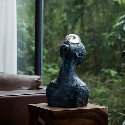 Abstract sculpture with a reflective ball on a wooden surface, blurred greenery in the background
